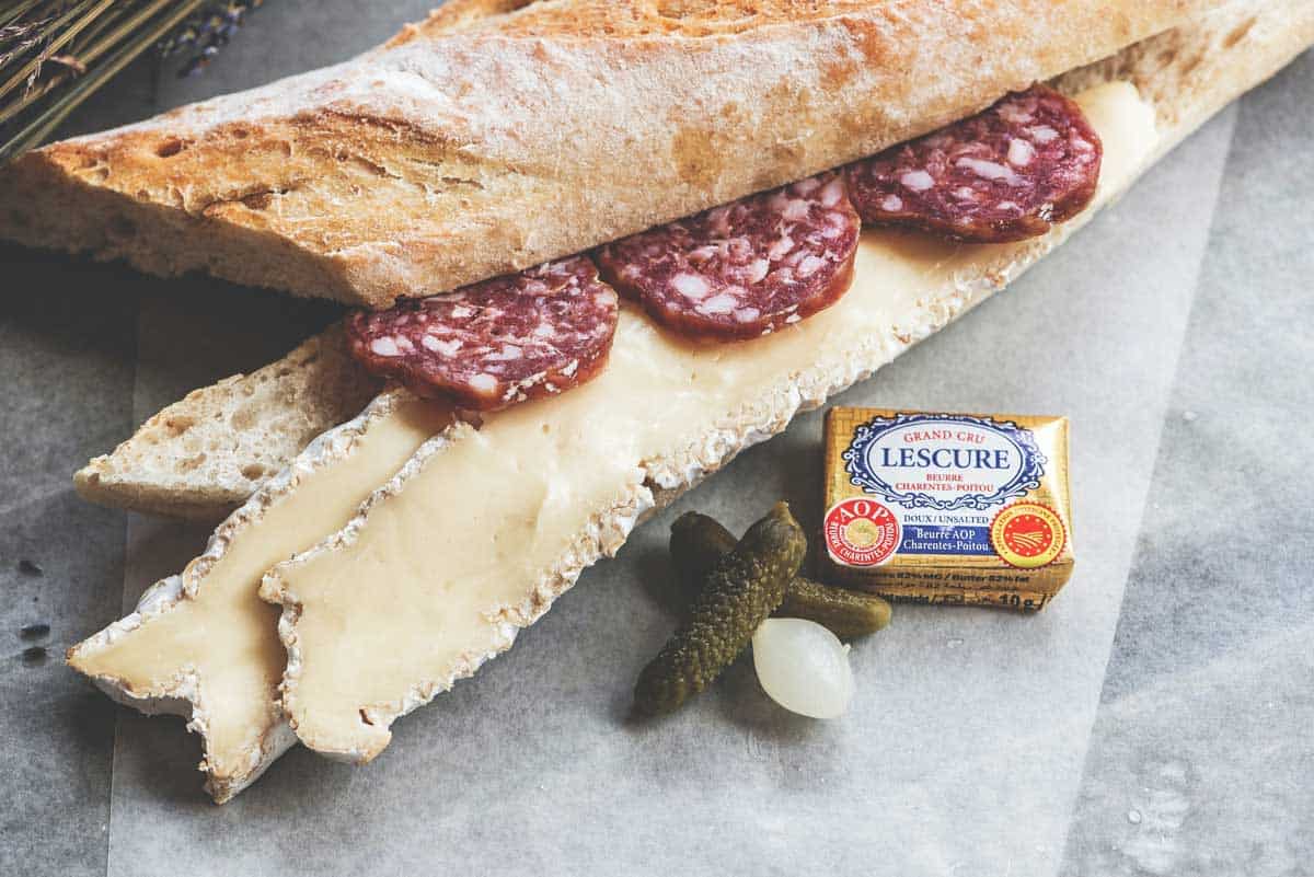 A French baguette with brie and salami with pickles and block of butter next to it. 