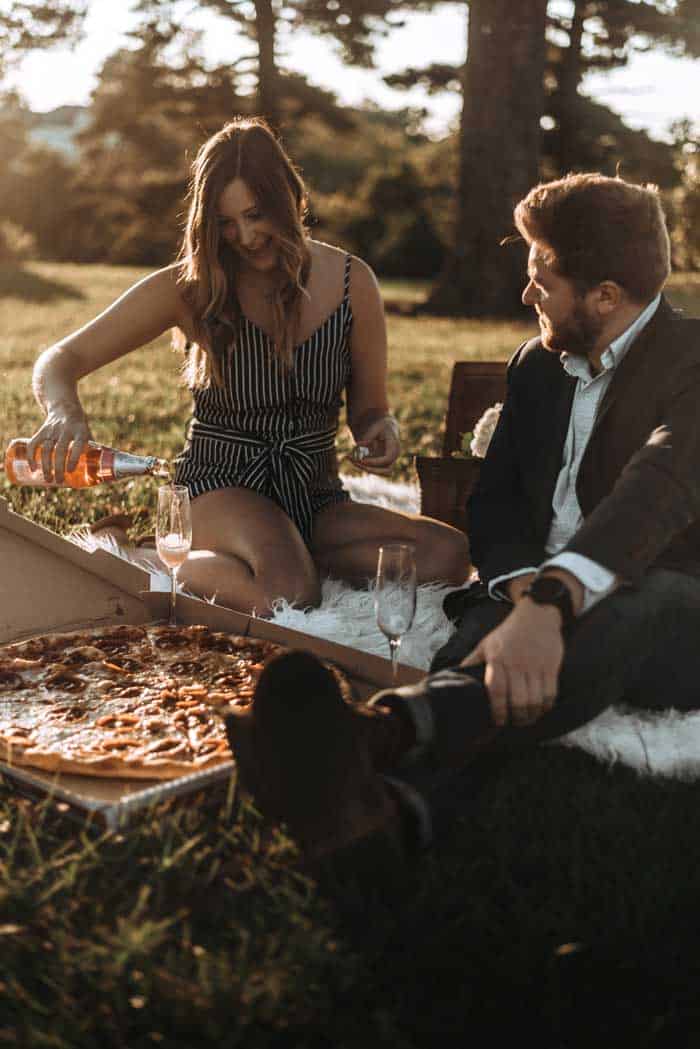 Couple enjoying champagne and pizza on a rug in the park. 