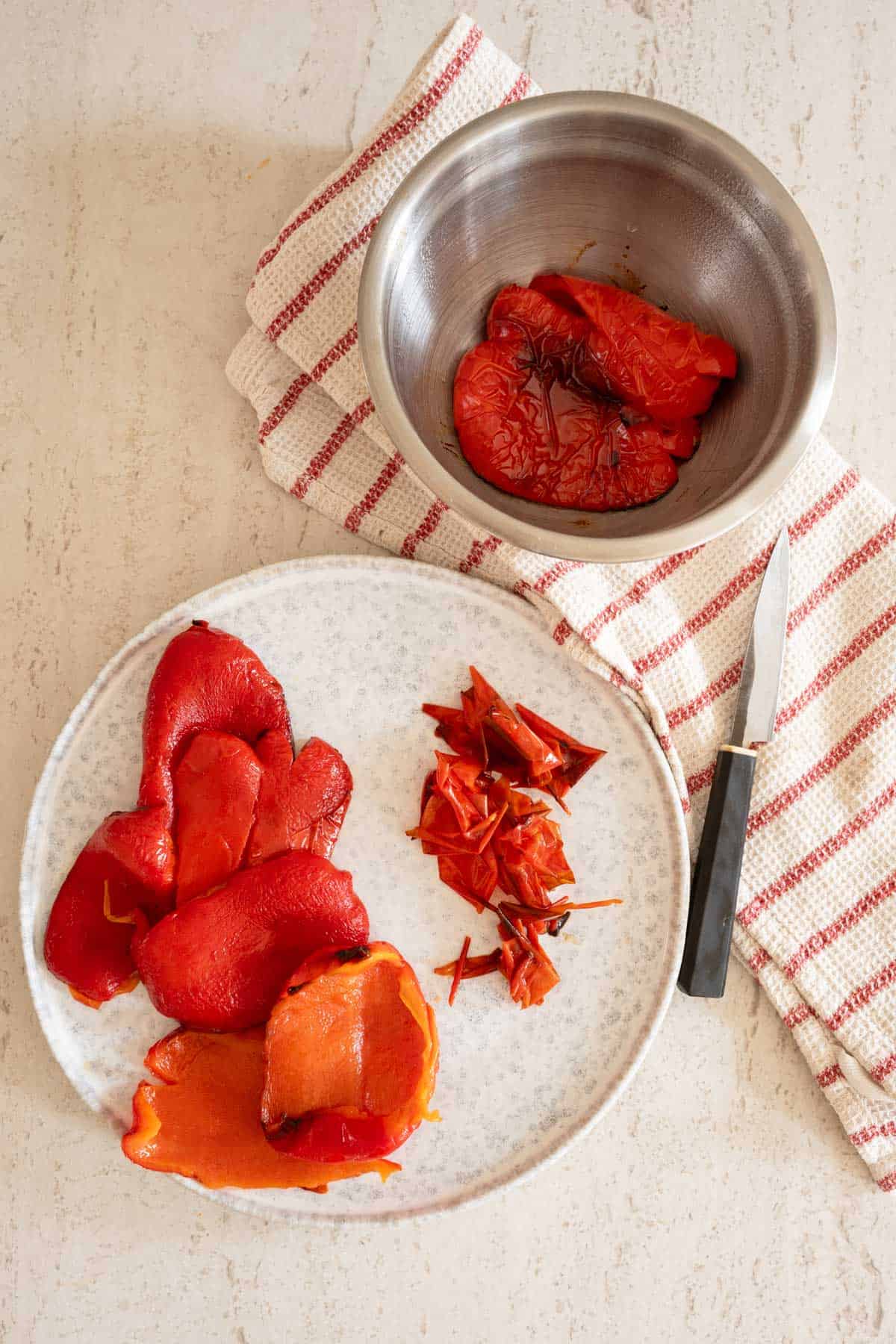 Roasted red peppers on a plate with thier skins to the side and a bowl of additional peppers on a kitchen counter.