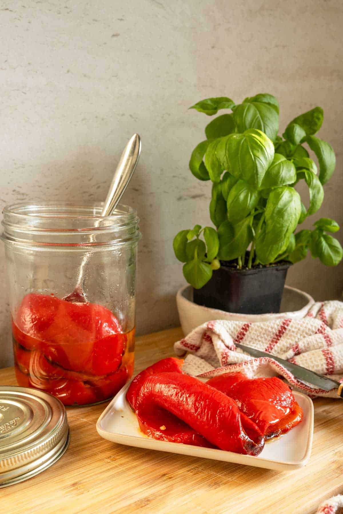 A jar of roasted red peppers with some on a plate and a basil plant beside on a wooden surface.