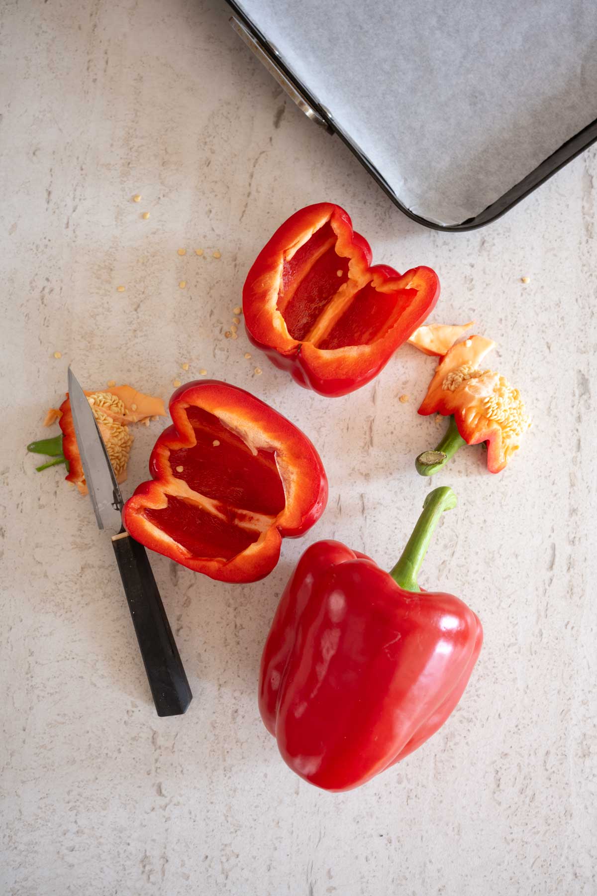 Red bell pepper cut in half with seeds on a kitchen counter alongside a knife.