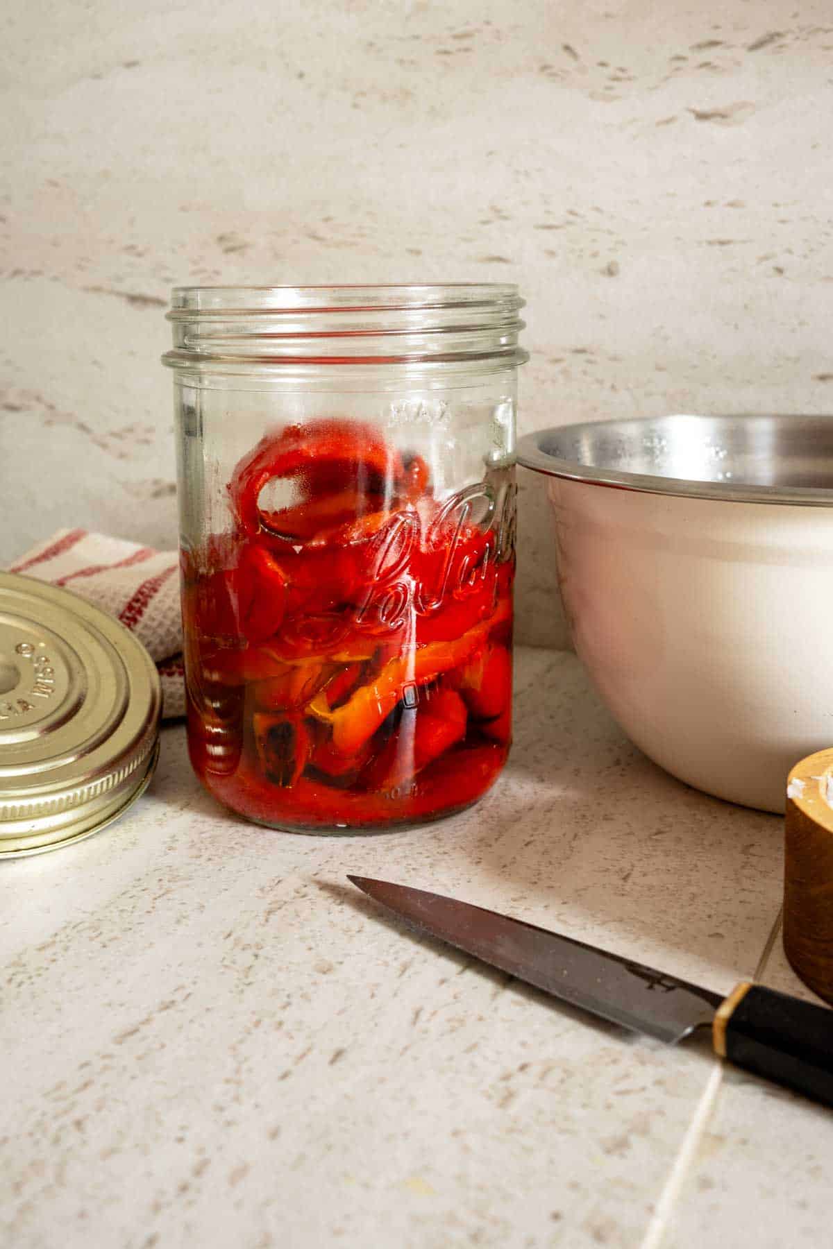 A jar filled with sliced roasted red peppers on a kitchen countertop, with a knife and an empty bowl nearby.