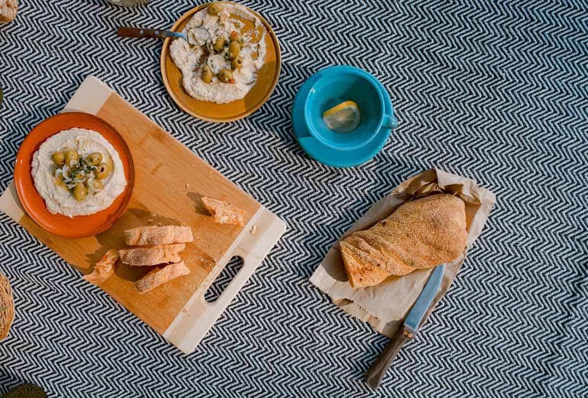 Blue picnic blanket with plates of hummus and a loaf of bread.