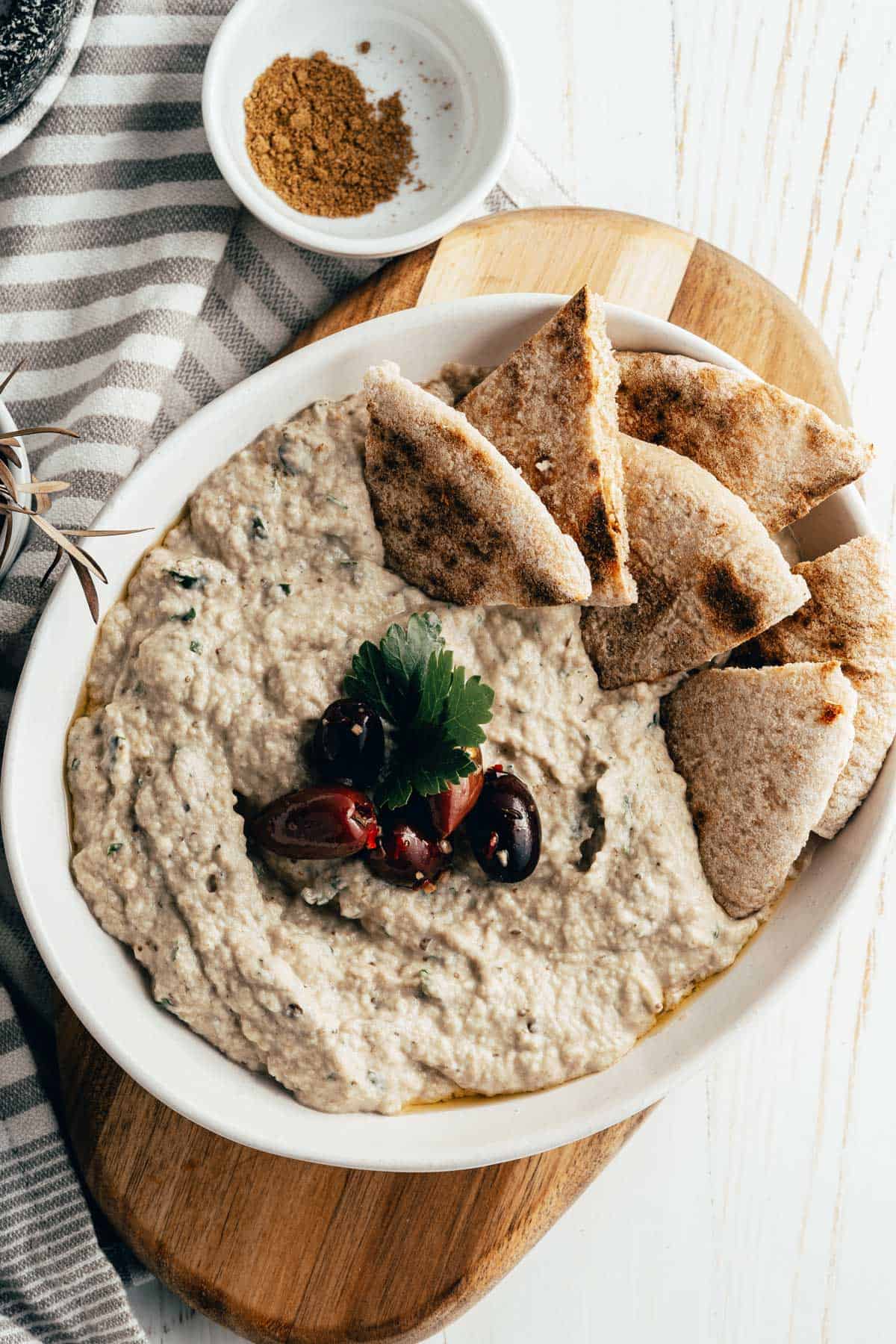 A bowl of baba ganoush eggplant dip garnished with herbs and olives, served with pita bread slices, is placed on a wooden cutting board. 