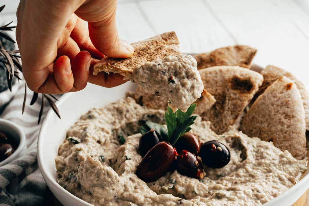 A hand dips pita bread into a bowl of creamy dip garnished with olives and herbs. 