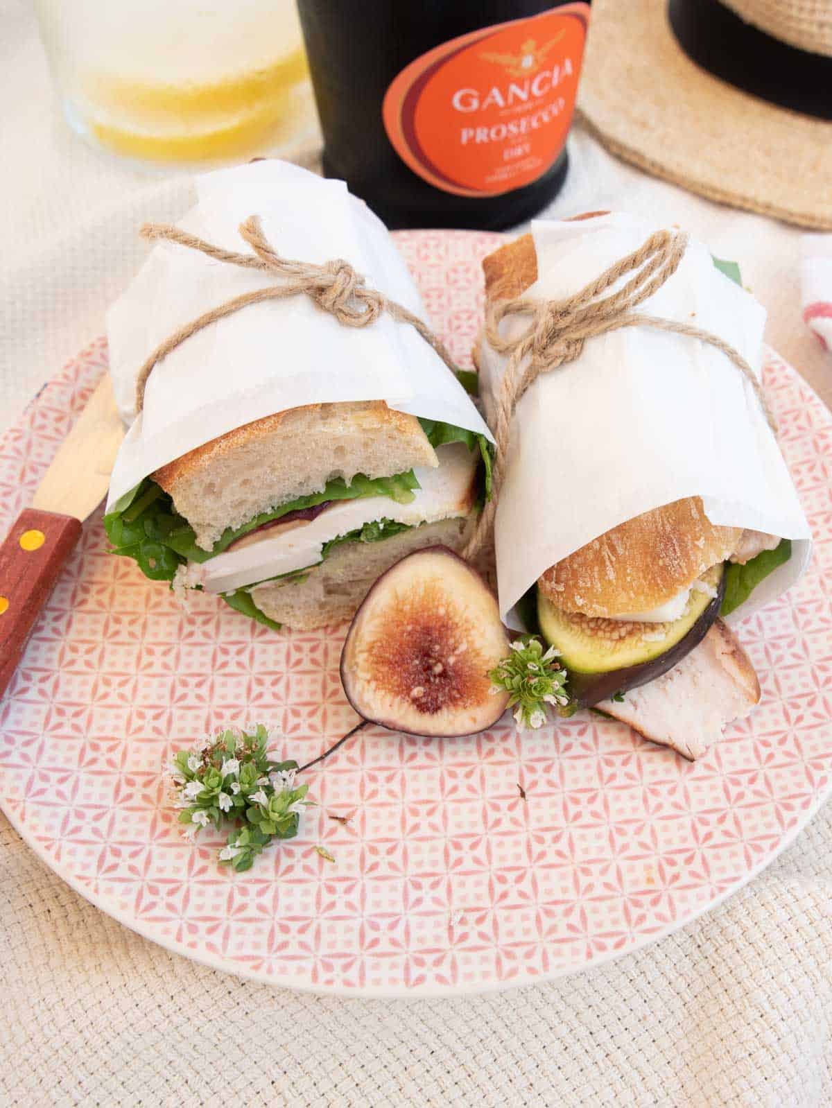 Two sandwiches wrapped in paper and tied with string on a plate next to a bottle of wine.