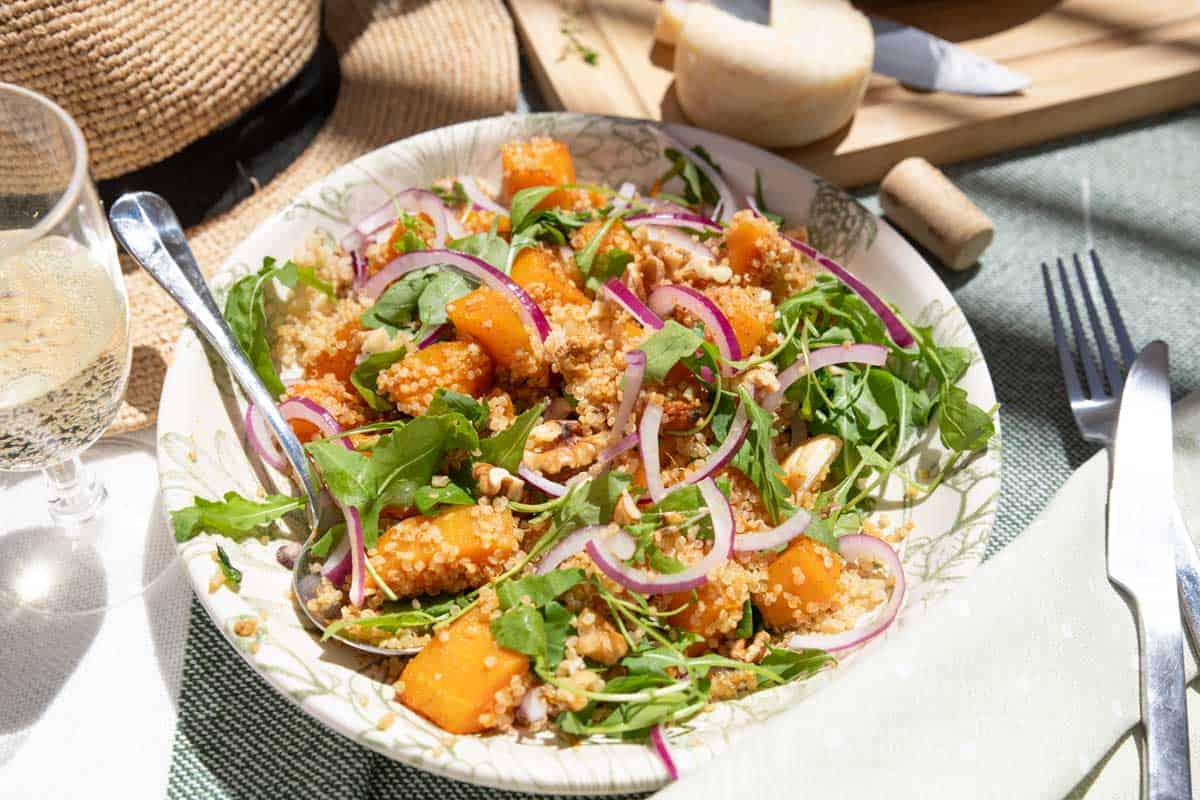 A plate of quinoa salad with chunks of roasted butternut squash, arugula, sliced red onions, and nuts. A glass of white wine, fork, knife, cloth napkin, hat, and cheese board are in the background.