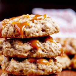 Close-up of a stack of three caramel-drizzled apple cookies on a wooden surface.