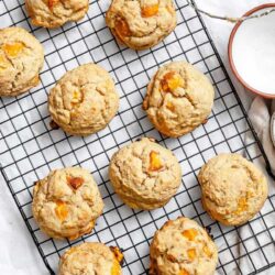 Nine apricot cookies cooling on a wire rack on a white surface, with a small bowl of white granulated sugar nearby.