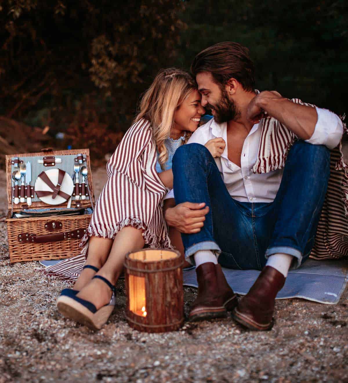 A couple is sitting on a picnic blanket cuddling with a picnic basket behind them.