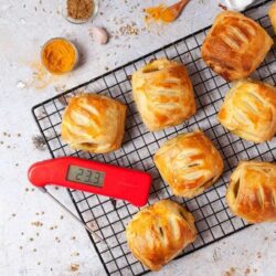 Seven golden-baked puff pastries on a cooling rack with a red digital thermometer displaying 23.3°C. Various spices and garlic cloves are scattered around.