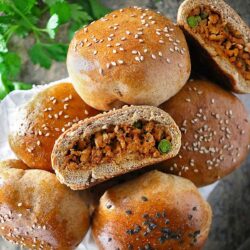 A close-up of several stuffed buns, some topped with sesame seeds. One bun is halved, revealing a filling of textured ground mixture with pieces of vegetables. Fresh herbs are in the background.