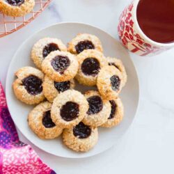 A plate of thumbprint cookies with dark jam centers, accompanied by a cup of tea in a red and white mug.