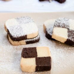 Square checkerboard cookies with alternating dark and light sections, dusted with powdered sugar, arranged on a beige surface.