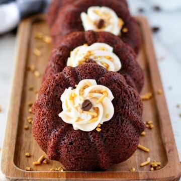 Three mini chocolate bundt cakes topped with vanilla frosting and sprinkled with golden and chocolate sprinkles are arranged on a wooden tray.