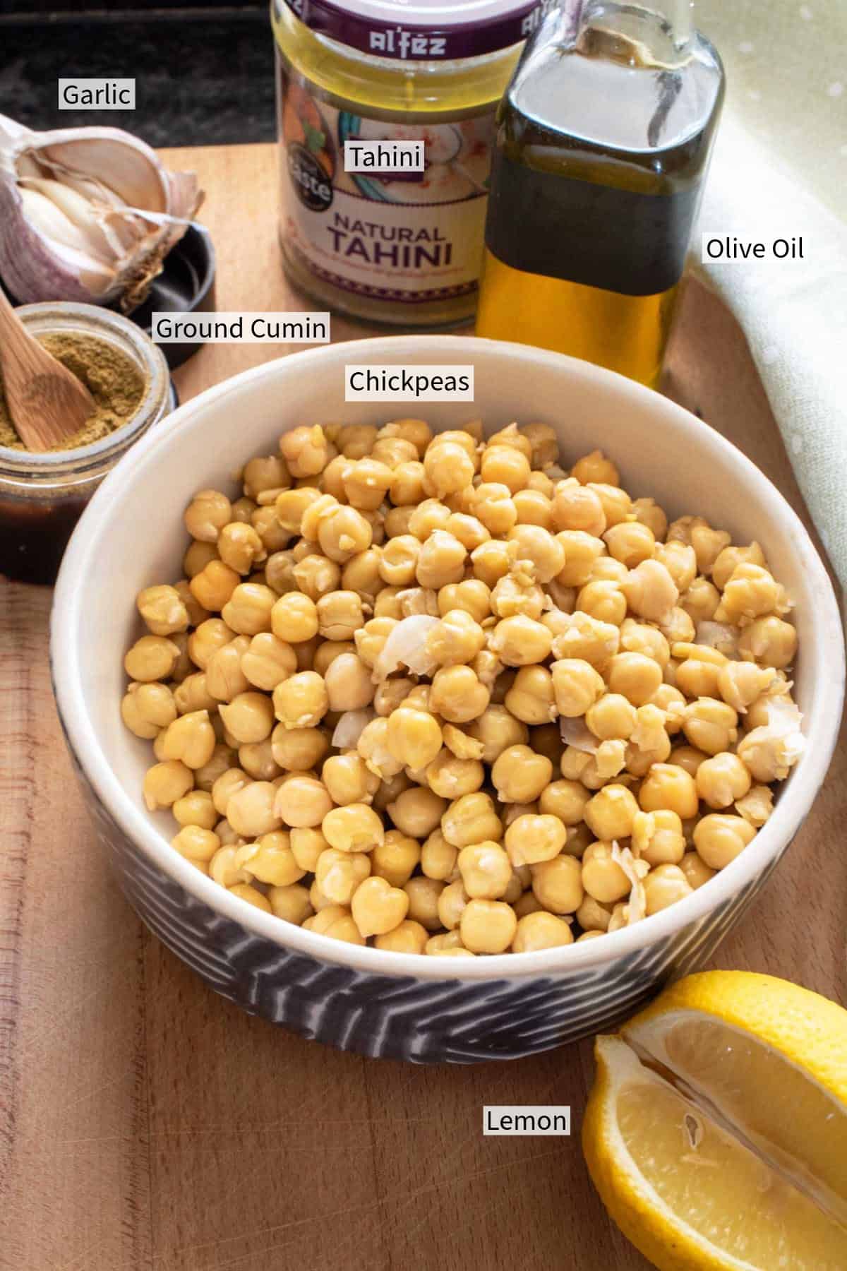 Ingredients for hummus on a wooden surface: a bowl of chickpeas, a bottle of olive oil, a jar of tahini, a sliced lemon, garlic, and ground cumin.