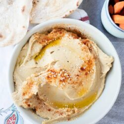 A bowl of hummus garnished with sesame seeds, paprika, and olive oil, next to pita bread and a small bowl of sliced carrots.