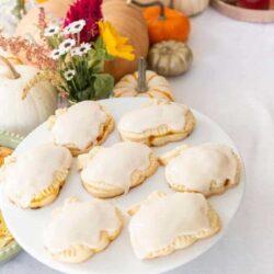 A white plate holding six frosted, pumpkin-shaped pastries is set on a table with fall decorations, including pumpkins and flowers, in the background.