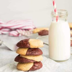 Stack of four marbled chocolate and vanilla cookies beside a bottle of milk with a striped straw on a light surface. Baking cooling racks with cookies are in the background.