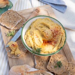 A bowl of hummus topped with sesame seeds and spices, surrounded by slices of whole-grain bread on a wooden board, with a knife placed beside the bowl.