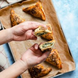 Hands holding a split triangular pastry with a spinach and cheese filling above a tray of similar golden-brown pastries.