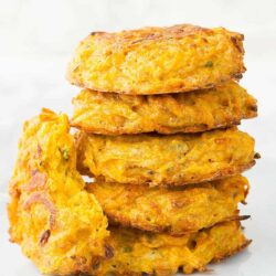 A stack of five golden-brown, crispy, homemade squash fritters with one partially broken on the side.