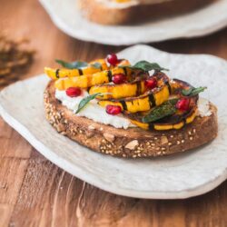 A slice of multigrain bread topped with roasted squash, pomegranate seeds, fresh herbs, and whipped cheese on a white plate.