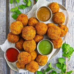 A plate of crispy, golden-brown fritters served with three different dipping sauces: one red, one green, and one brown, garnished with fresh coriander leaves.