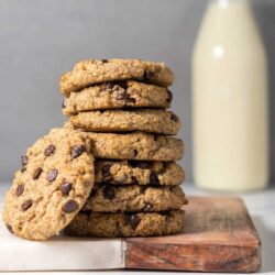 A stack of chocolate chip cookies is placed on a wooden board with a bottle of milk in the background.