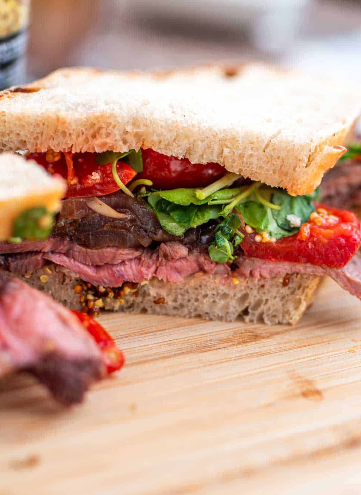 A close-up of a roast beef sandwich with tomatoes, greens, and mustard on whole-grain bread.