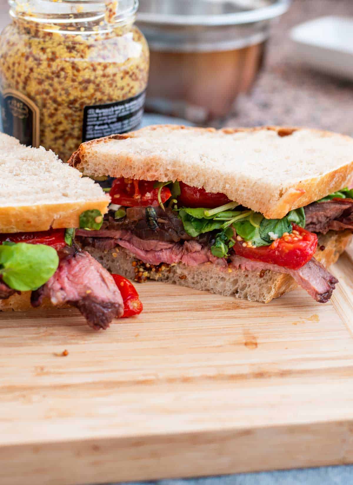 A steak sandwich with arugula and roasted peppers on a wooden cutting board, with a jar of mustard in the background.