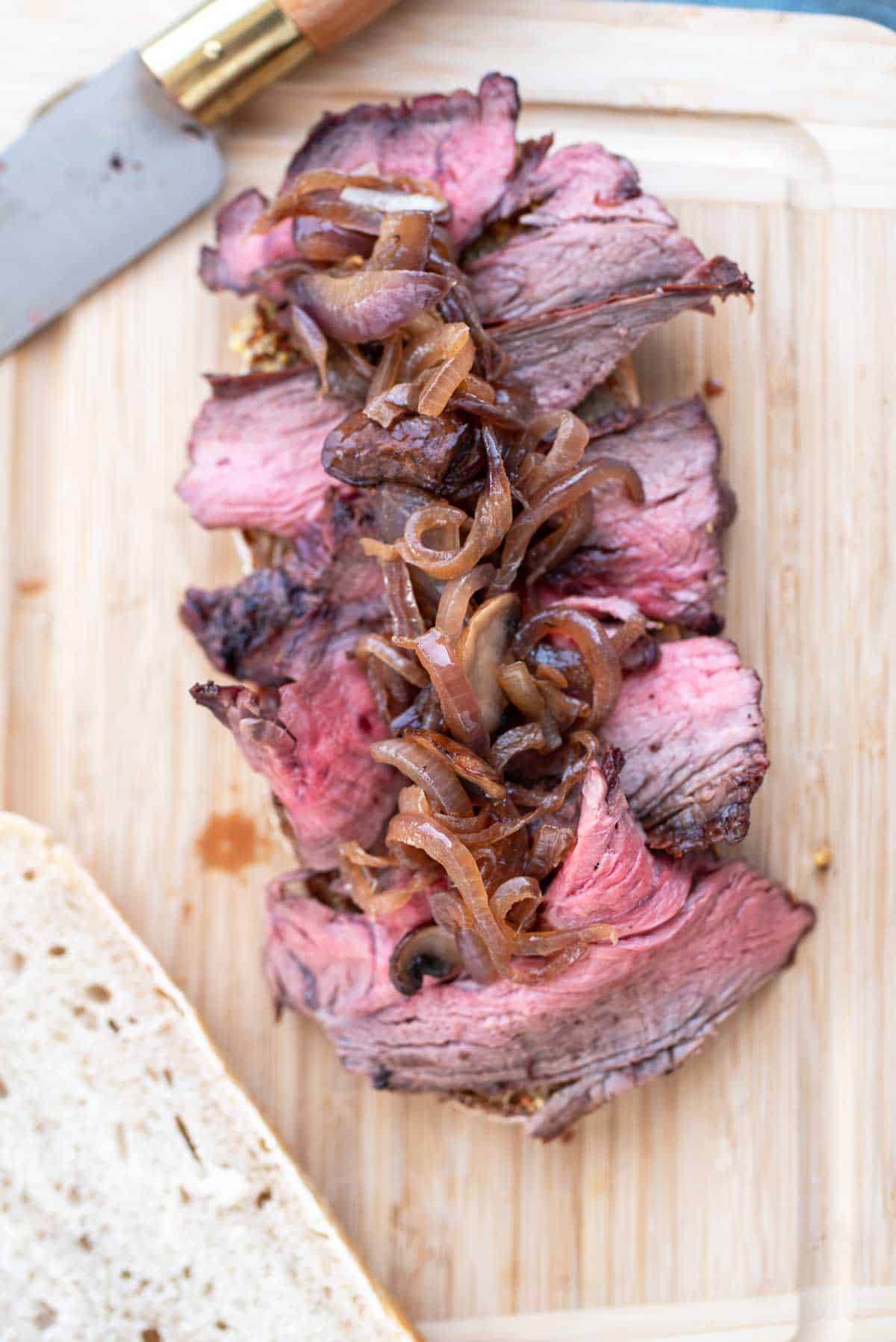 Sliced medium-rare beef on a slice of bread topped with caramelized onions on a wooden cutting board.