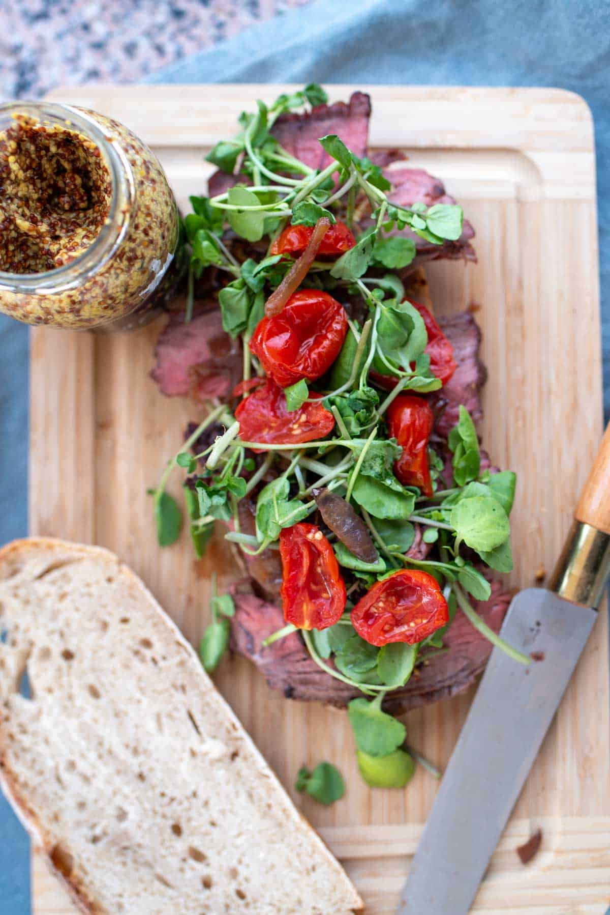 Open-faced roast beef sandwich with arugula and cherry tomatoes on a wooden cutting board.