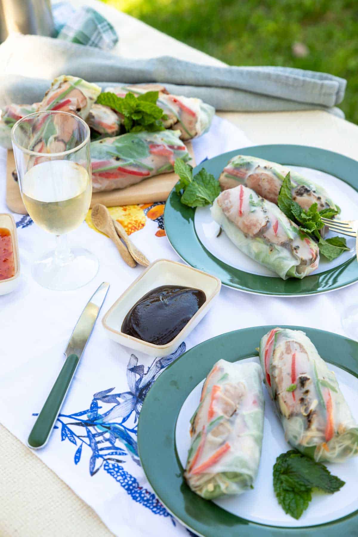 A picnic table with plates of summer rolls, asian sauces and glasses of wine.