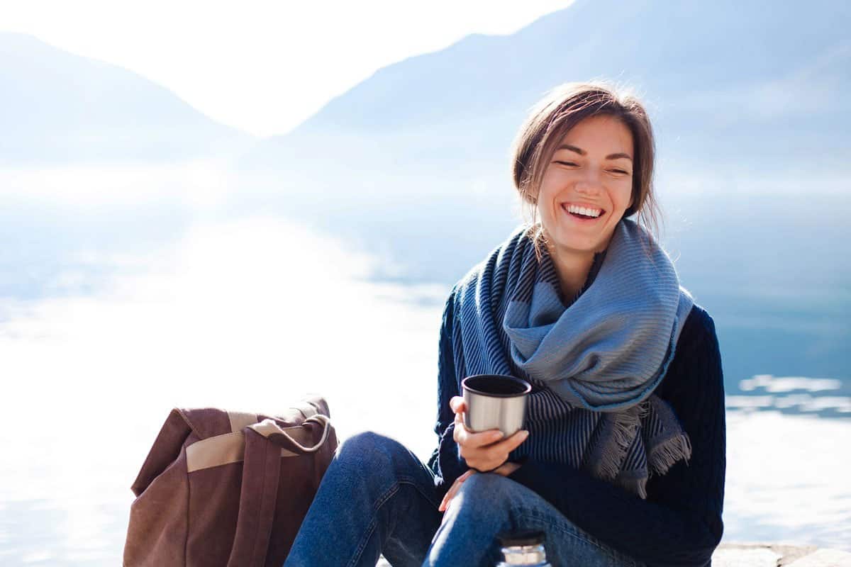 Smiling lady holding thermos cup in front of lake in winter. 
