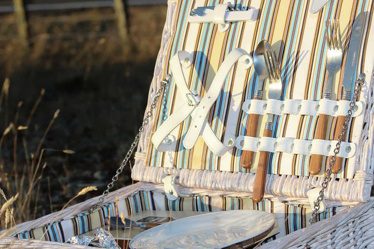 Close up of open white wicker picnic basket in a field with cutlery in the place holders inside the lid. 