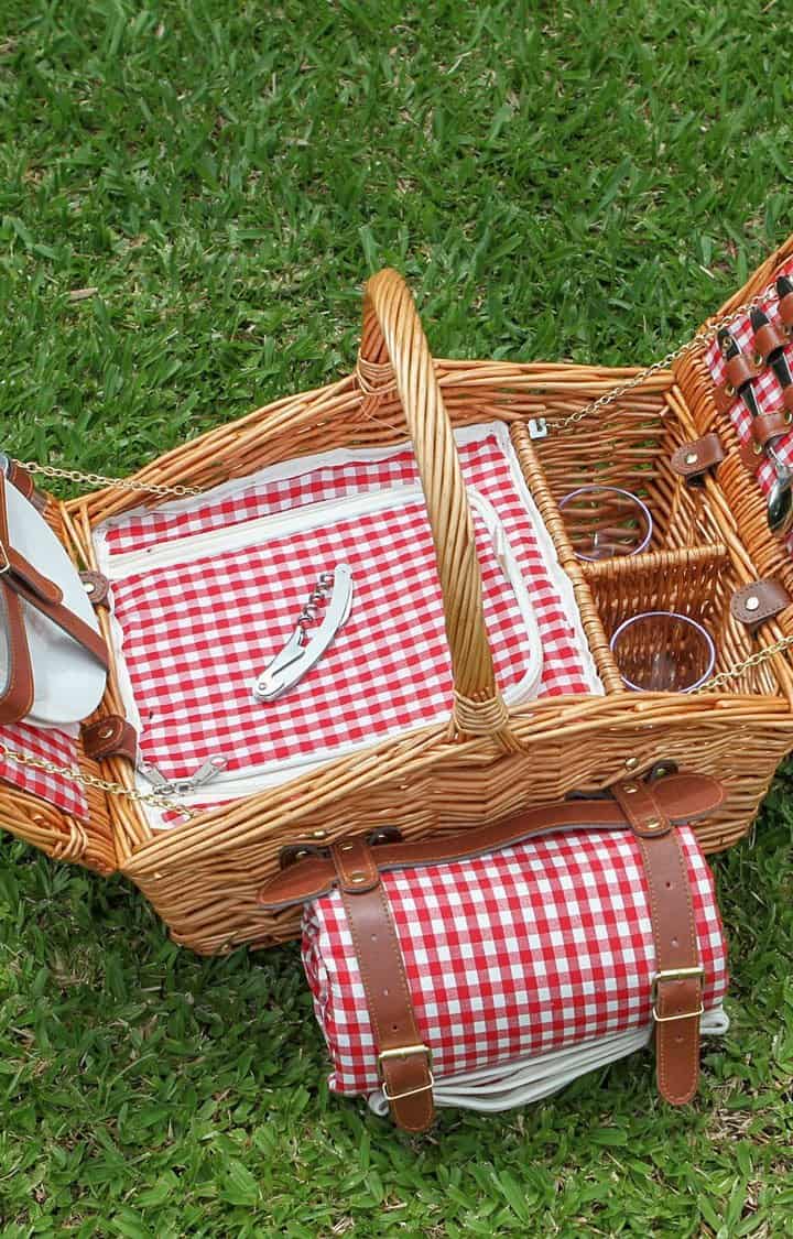 Traditional brown wicker picnic basket on green grass with rolled red check blanket and matching insulated insert.