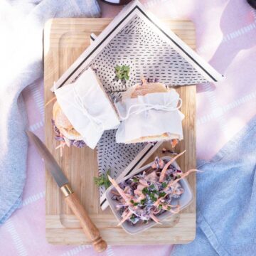A picnic scene with two sandwiches wrapped in cloth, coleslaw on a plate, and a knife on a wooden cutting board, all atop a pink cloth.