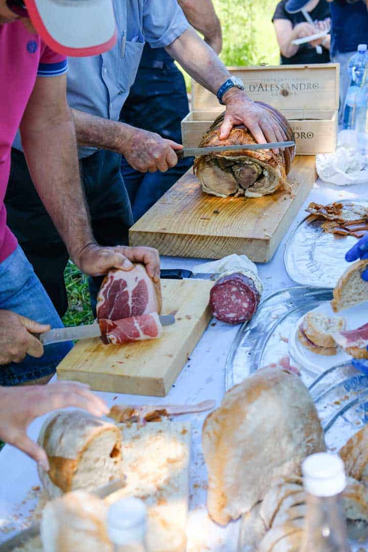 people slicing cold cuts on a picnic table for sandwiches. 