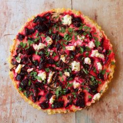 A vegetable tart with a golden crust, topped with pieces of beets, feta cheese, caramelized onions, and fresh herbs, shown on a rustic wooden surface.