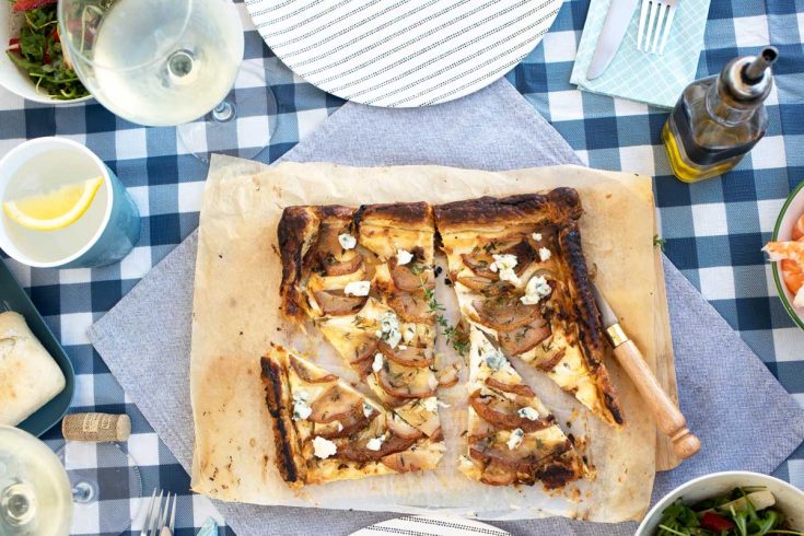 Slices of Pear and ricotta tart on baking paper laid on a blue check picnic table cloth.