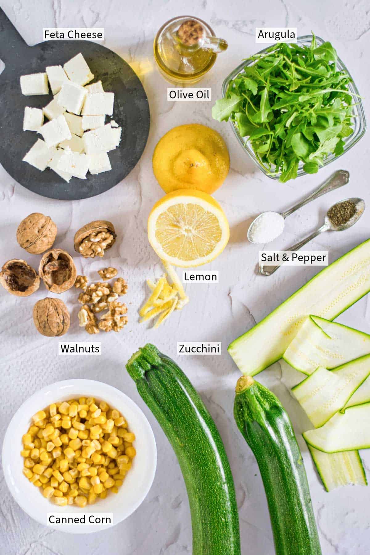 Ingredients for a zucchini and arugula salad with feta cheese, walnuts, canned corn, and a lemon olive oil dressing.