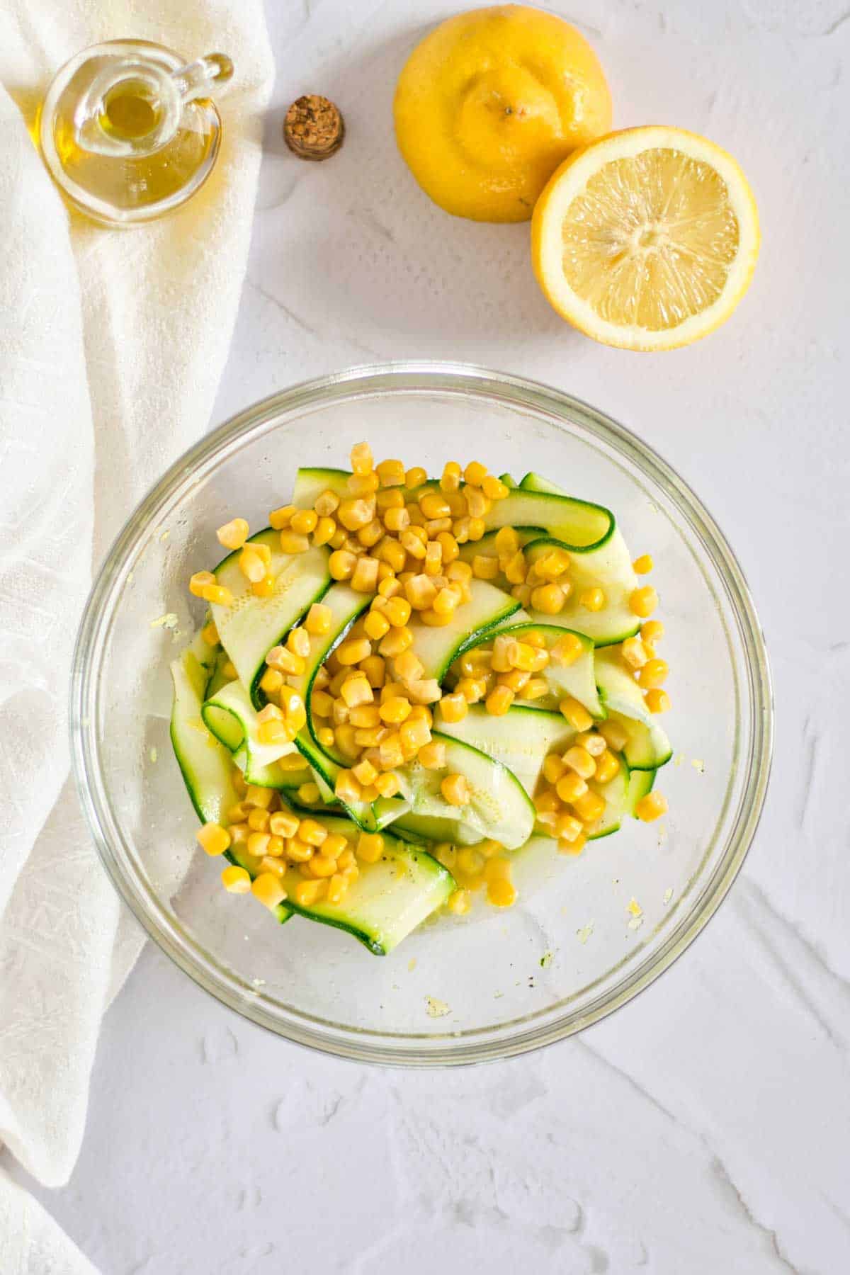 A bowl of zucchini ribbons and corn salad with lemon and a bottle of olive oil on a white surface.