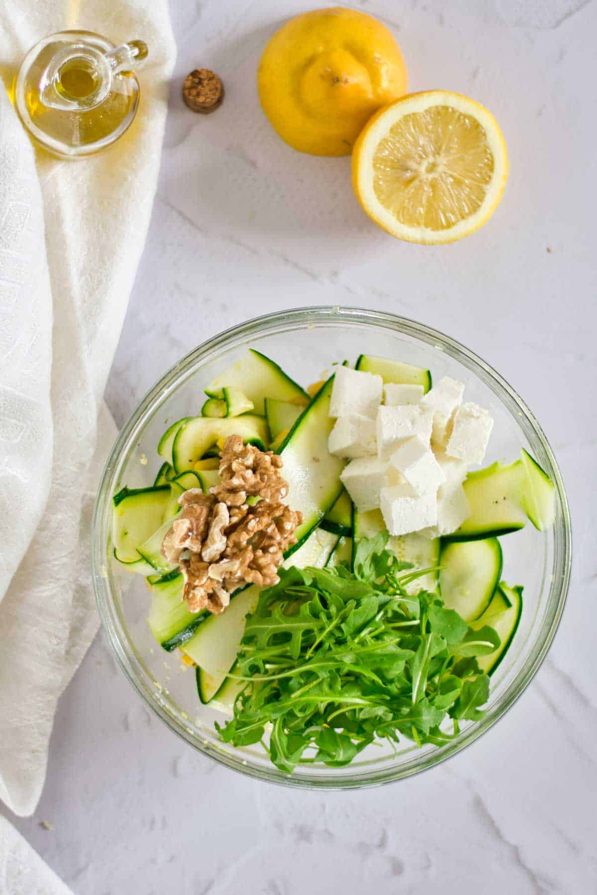 A bowl of arugula, cucumber ribbons, feta cheese, and walnuts with lemons and olive oil on the side on a white surface.