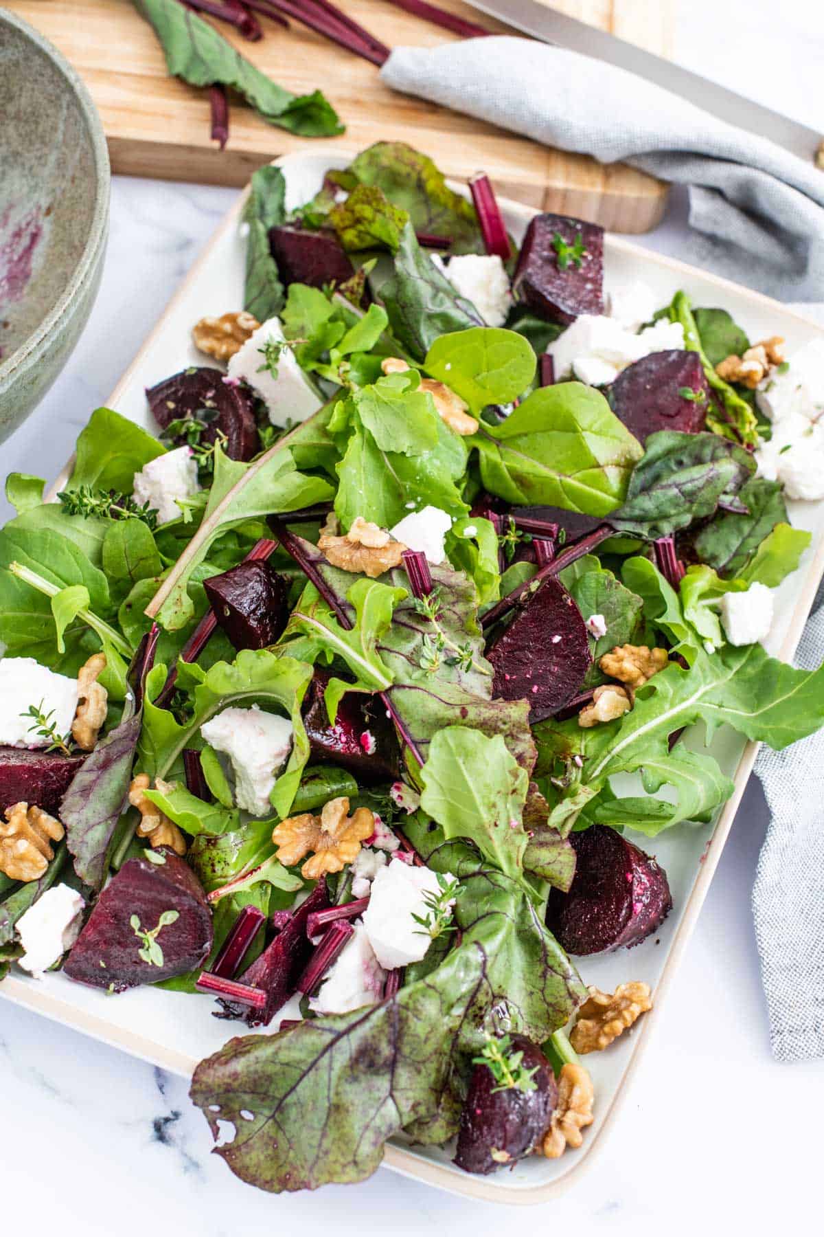 A fresh beetroot salad with mixed greens, walnuts, and fetacheese on a rectangular plate.