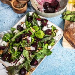 A vibrant beetroot salad with feta cheese, mixed greens, and nuts, on a blue textured background.