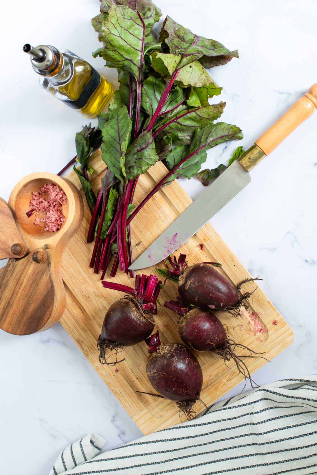 Fresh beetroots with leaves on a cutting board alongside a kitchen knife, olive oil, and salt.