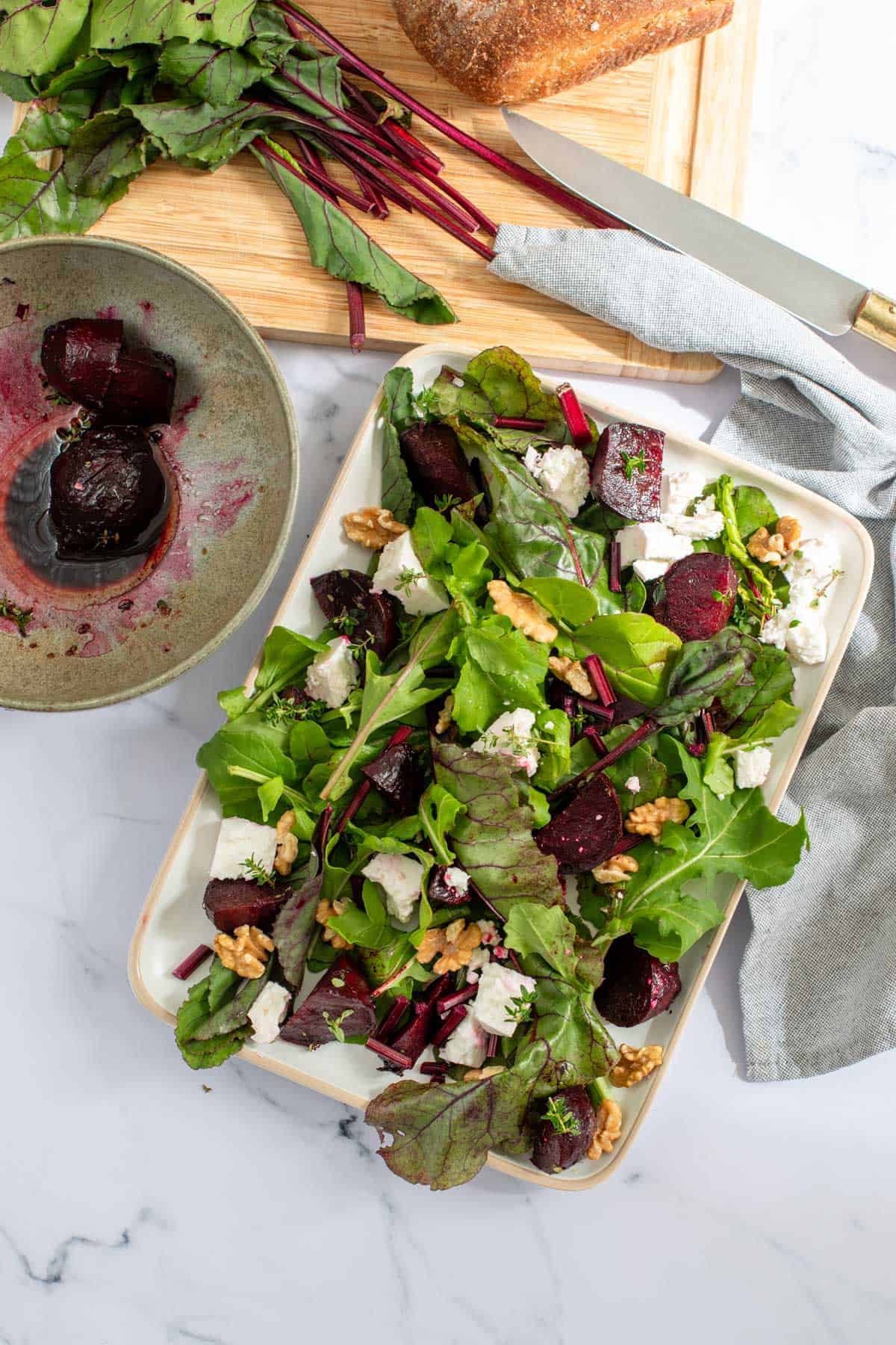 Fresh beetroot salad with mixed greens, nuts, and cheese, served on a rectangular platter, with ingredients nearby on a marble countertop.