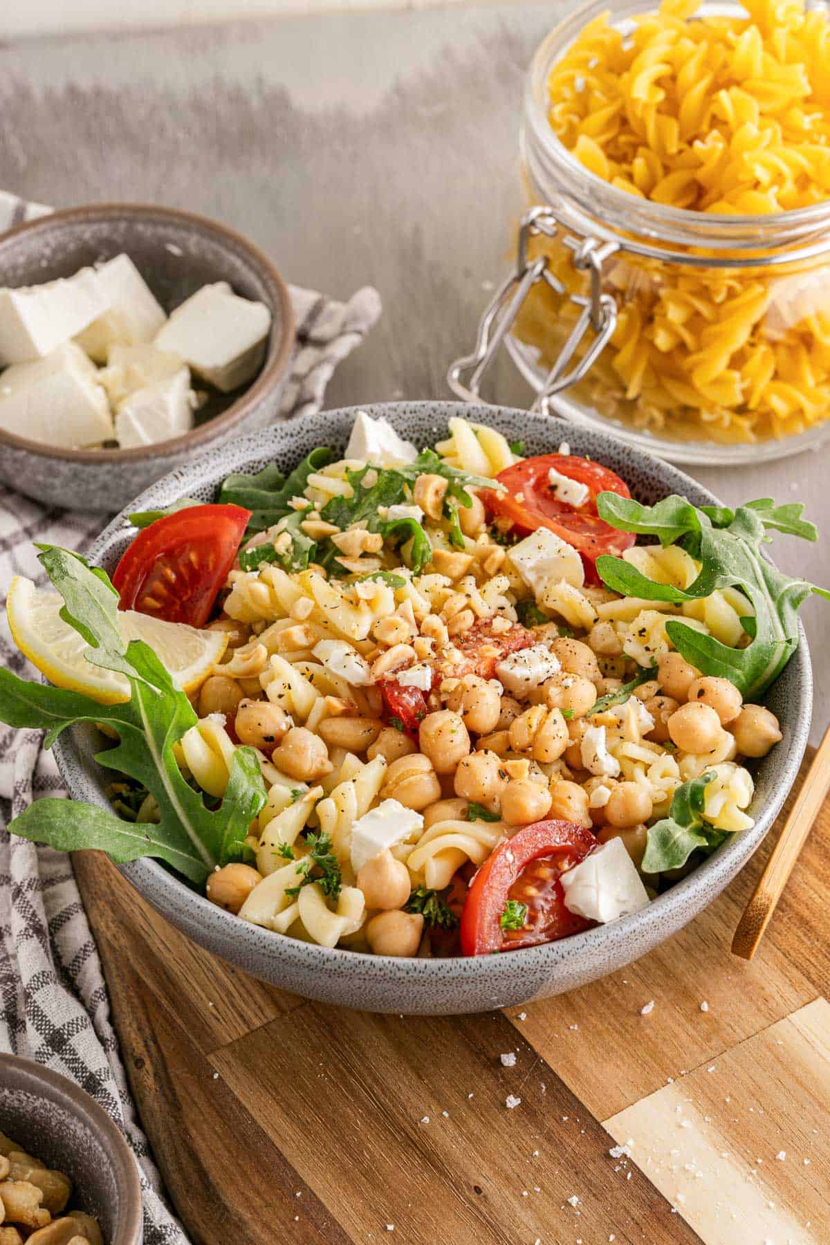 A bowl of chickpea pasta salad with tomatoes, arugula, pasta, and cheese on a wooden table, alongside ingredients.