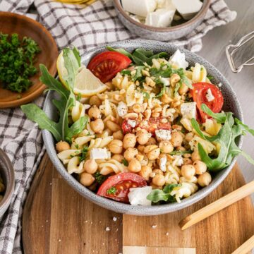 A bowl of chickpea salad with tomatoes, cheese, and arugula on a wooden table, garnished with herbs and lemon.